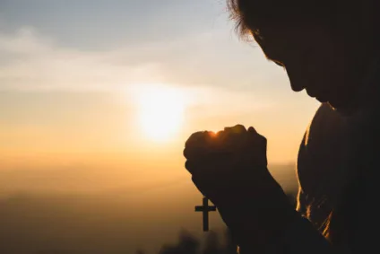 Silhouette of young  human hands praying with a  cross at sunrise, Christian Religion concept background.